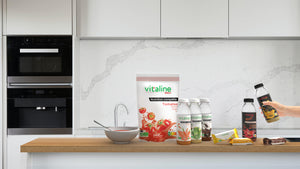 Vitaline Tomates en sachet avec un bol trois bouteilles de Vitaline Carottes Légumes Verts et Cacao avec barre Bananes Carottes Cacao et bouteille Vitaline Catalyst Ignite et Focus posés sur un plan de travail en bois dans une cuisine